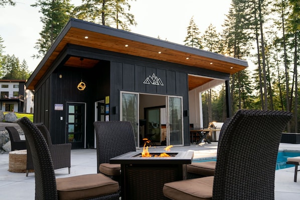 A modern outdoor seating area with a fire feature, surrounded by a pool and forested background.