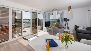 Una sala de estar moderna con un gran ventanal que da a un balcón, un comedor y una zona de estar con televisión y flores.