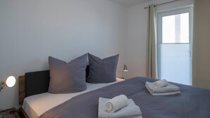 Una cama bien tendida con almohadas, una lámpara de noche y una ventana con cortinas.
