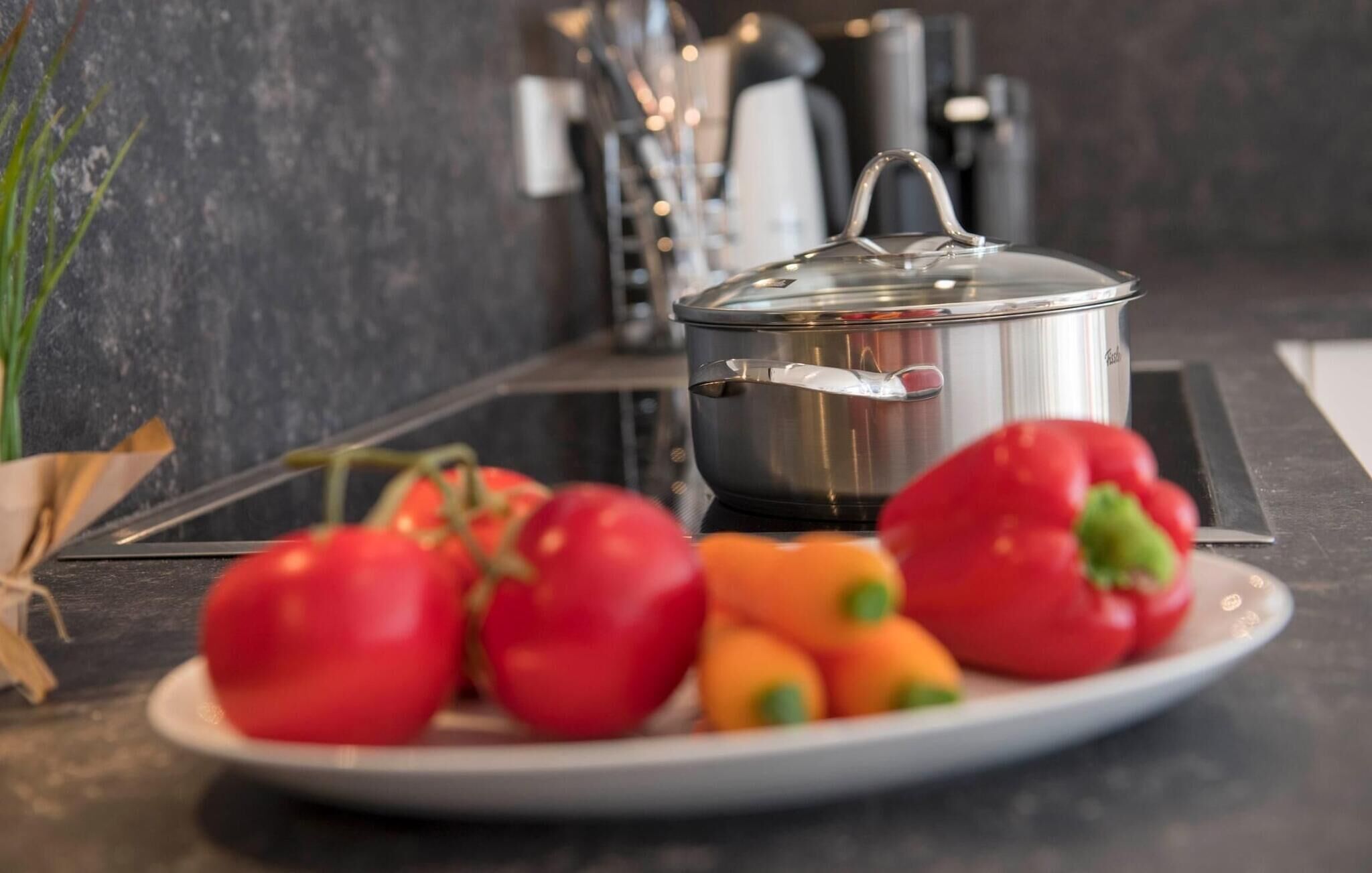 A stainless steel pot on a stove with a lid, surrounded by red and orange bell peppers on a white plate.