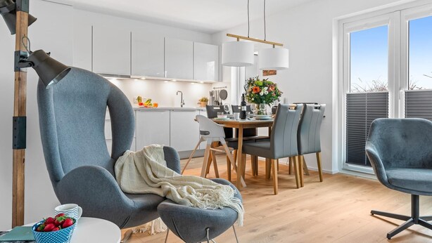 A modern kitchen with white cabinets, a dining area with a wooden table and chairs, and a large window with blinds.