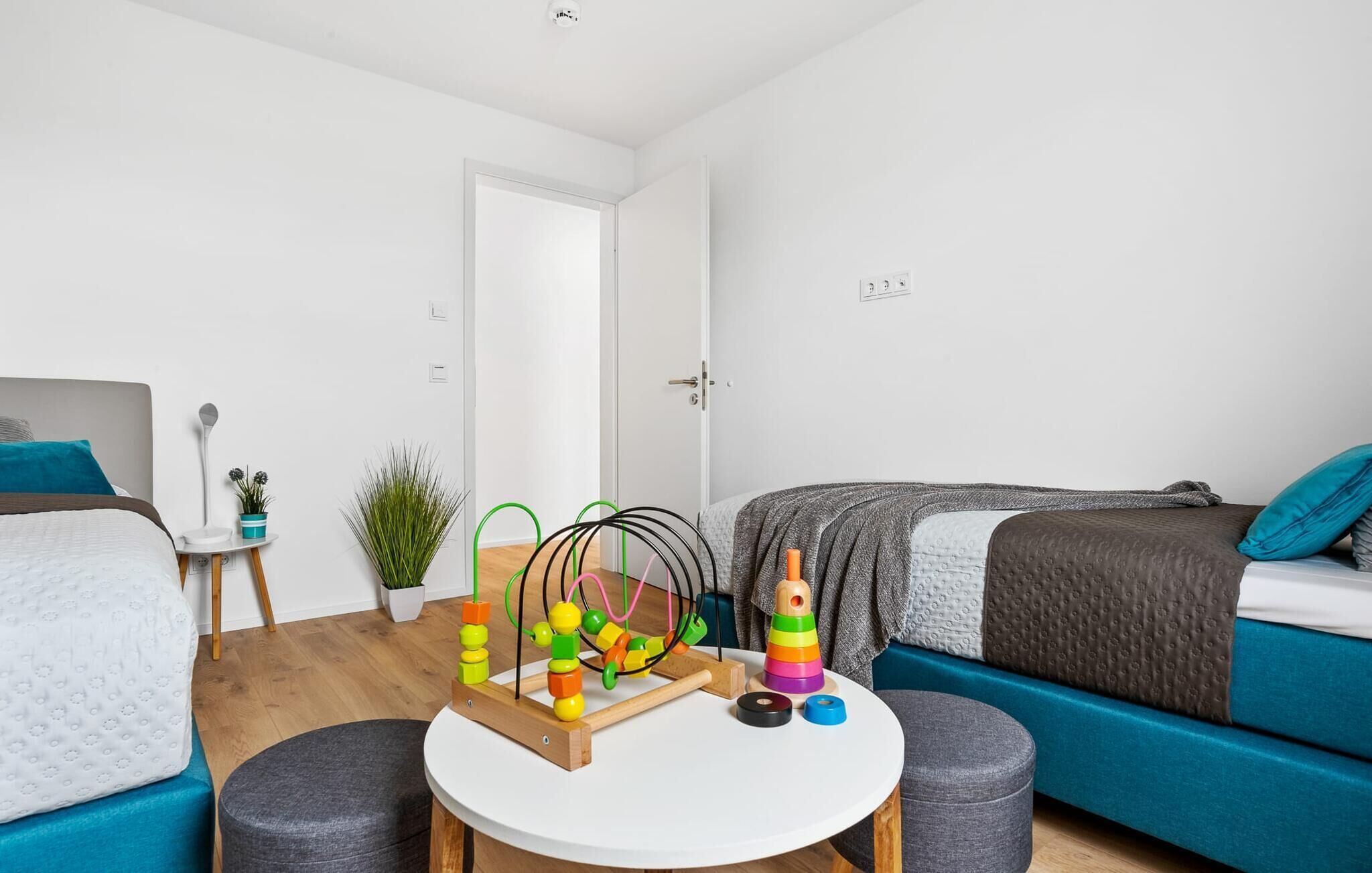 A modern, well-lit bedroom with a bed, a small round table with toys, a potted plant, and a blue sofa.
