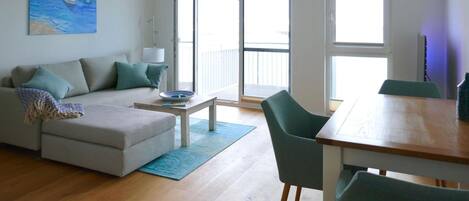 A modern living room with a sofa, a dining table, and a chair. There is a painting on the wall, a lamp, and a window with a view.