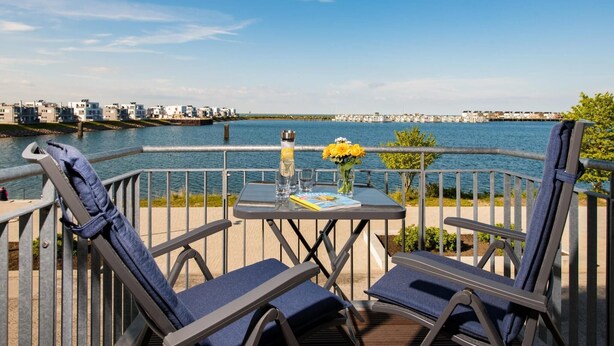 Eine Terrasse mit zwei Liegestühlen, einem Tisch mit einem Buch und einer Vase mit gelben Blumen, mit Blick auf eine städtische Skyline am Wasser.