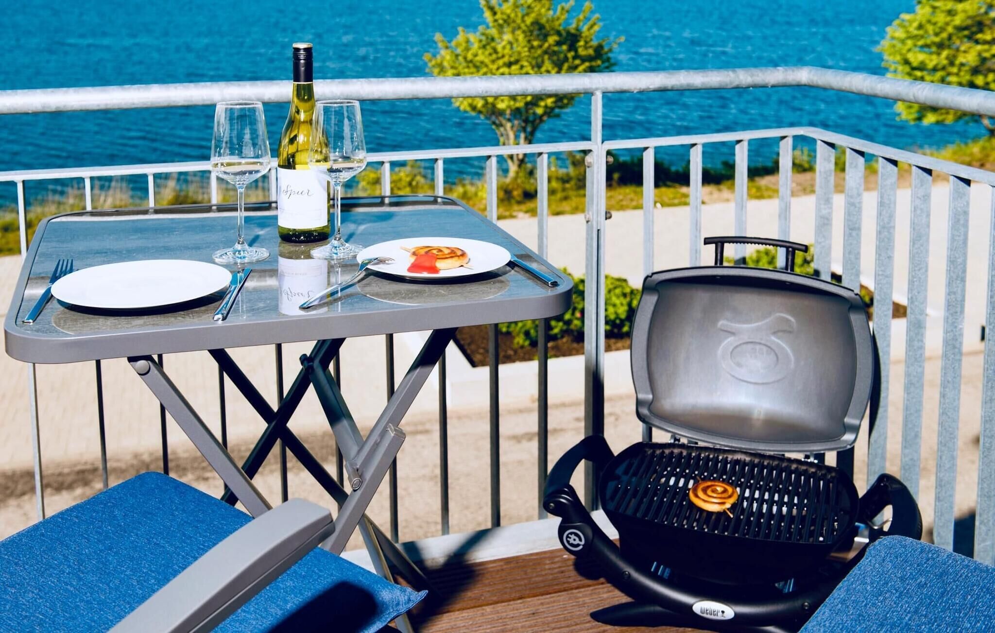 A table with a bottle of wine, glasses, and plates, a grill, and blue chairs on a balcony with a view of the sea.