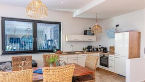 A modern kitchen with white cabinets, a wooden dining table, wicker chairs, and a large window with a view of a cityscape.