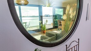 A circular window with a view of a balcony area featuring wicker furniture, a potted plant, and a view of a body of water.