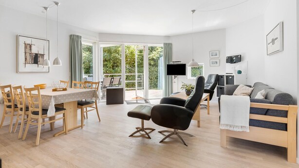 A modern living room with a dining area, wooden furniture, and a large sliding glass door leading to a balcony. - Gudhjem Søpark Lejl. 25 (Gudhjem)
