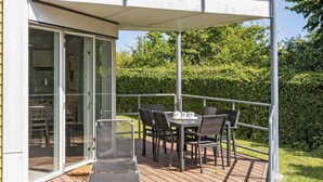 A covered outdoor seating area with a table and chairs, a glass door leading to a room, and a hedge in the background. - Gudhjem Søpark Lejl. 25 (Gudhjem)