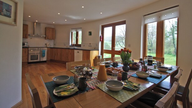 A modern kitchen with wooden cabinets, a dining table set with various dishes, and large windows offering a view of the outdoors. - 4 bedroom accommodation in Cilycwm, Llandovery (Cilycwm, Llandovery)