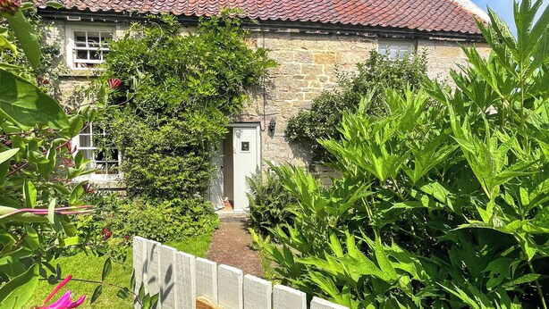 A stone house with a red-tiled roof, surrounded by lush greenery and a wooden fence with a sign reading "Pennywells."