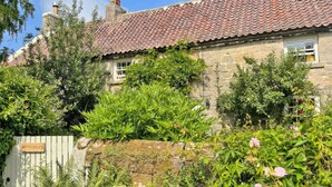 A stone house with a tiled roof, surrounded by lush greenery and flowering plants.