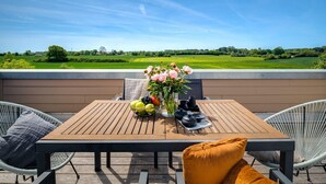 Ein Holztisch mit Blumen und Früchten in einer Vase, umgeben von Korbstühlen und Kissen, mit Blick auf eine grüne Landschaft.