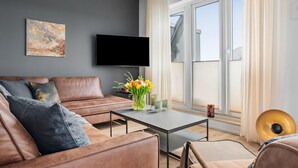 Ein modernes Wohnzimmer mit einer Ledersofa, einem Couchtisch mit Blumenvase, einem flachbildfernseher an der Wand und einem großen Fenster mit Vorhängen.