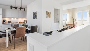 Eine moderne Küche mit weißen Schränken, ein Essbereich mit weißem Tisch und Stühlen sowie ein Blick ins Freie durch große Fenster.
