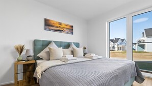 A bedroom with a large bed, a nightstand with a lamp, a painting on the wall, and a view of houses through the window.