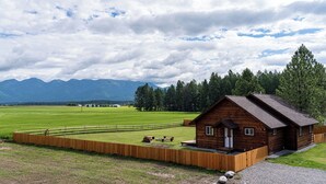 Una cabana de fusta amb un porxo, envoltada per una tanca de fusta, situada en un camp herbós amb muntanyes al fons.