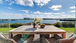 A wooden table with wicker chairs, a bottle, a glass, and a fruit bowl on a deck overlooking a body of water with buildings in the distance.