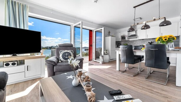 A modern living room with a large TV, a sofa with decorative pillows, a dining area with chairs, and a kitchen with a view of the sea.