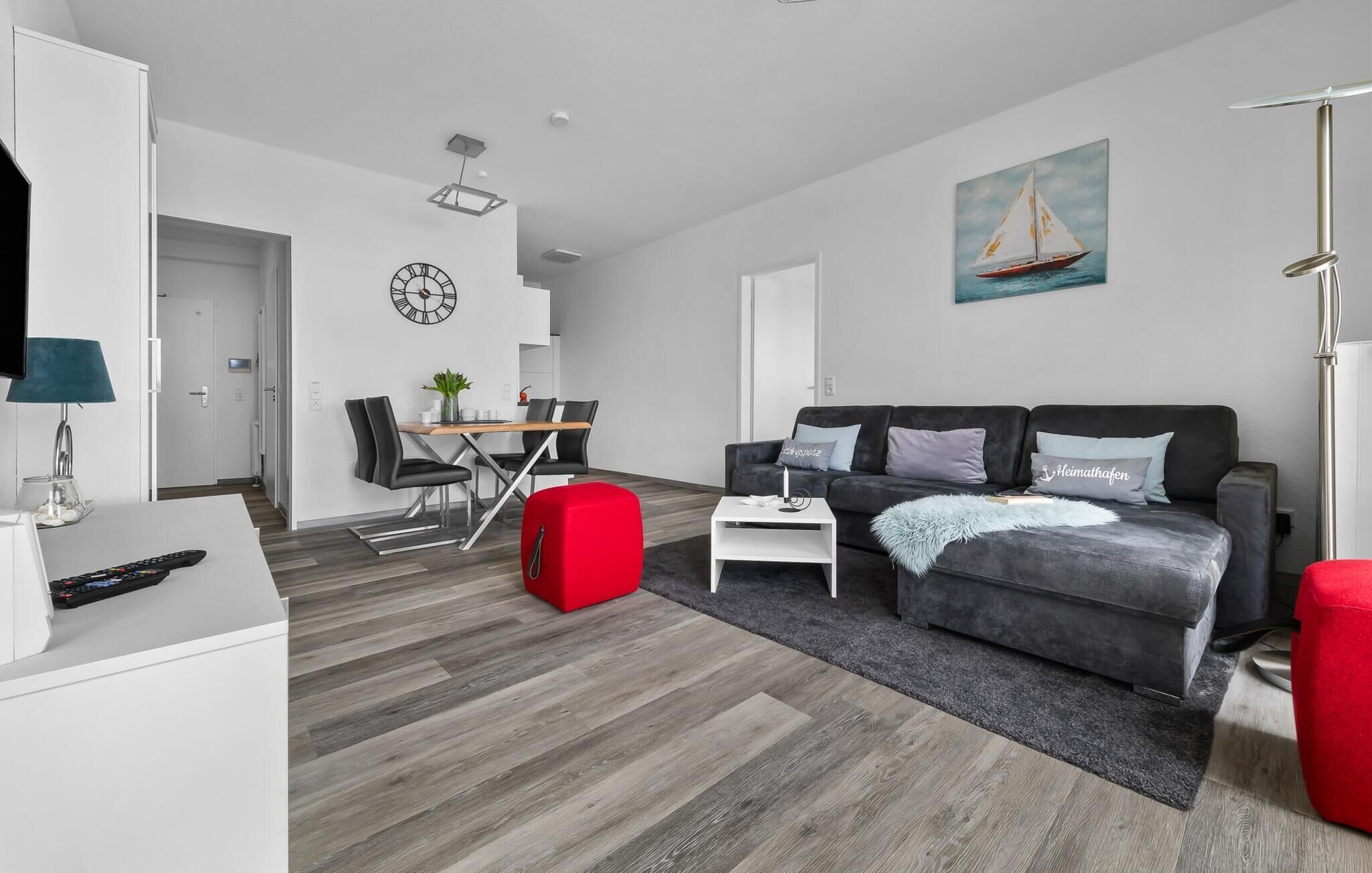 En moderne stue med en grå sofa, en rød skammel, et spiseområde med bord og stole, og et maleri af en sejlbåd på væggen.