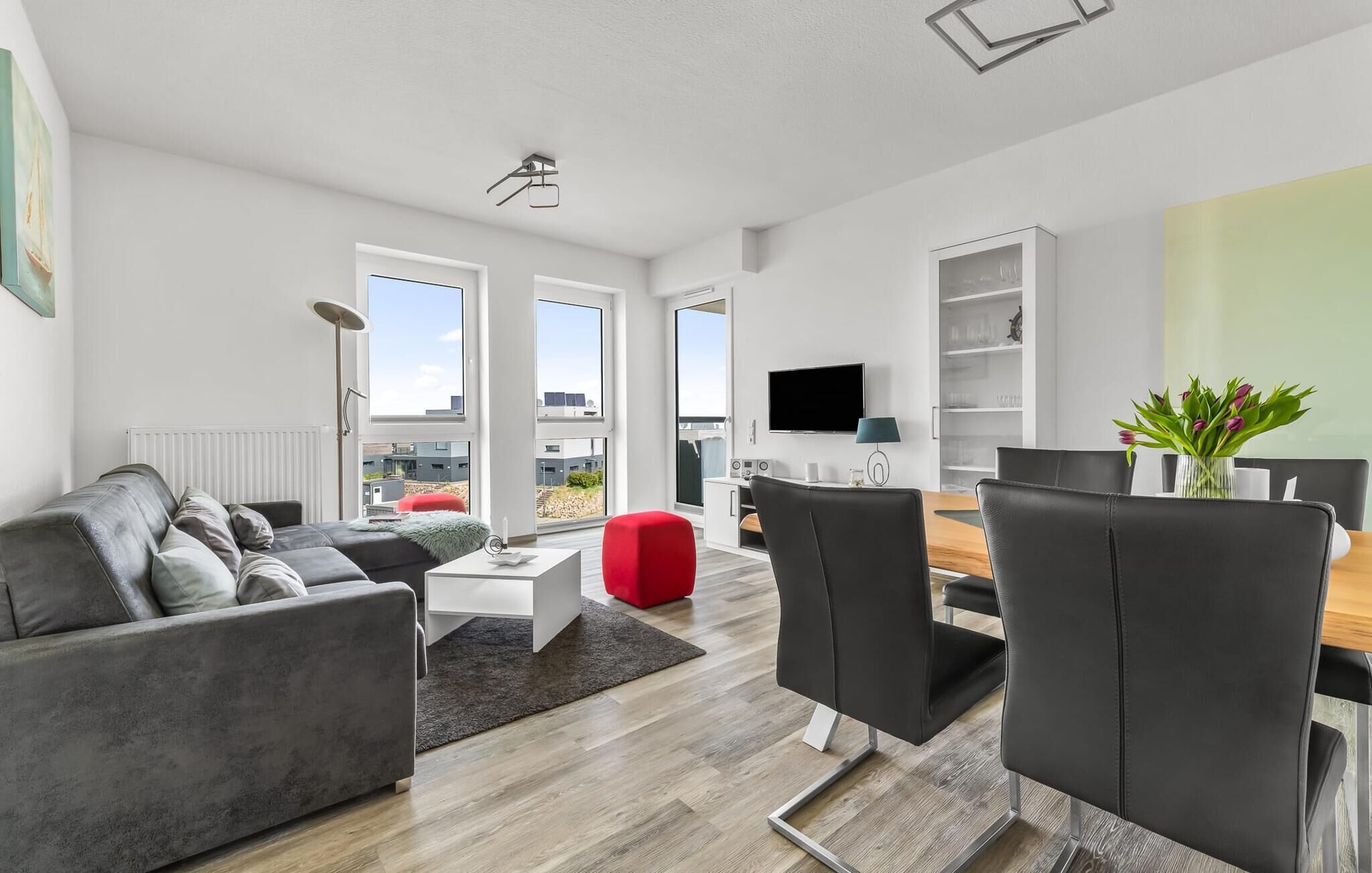 Una sala de estar moderna con un sofá gris, un otoman rojo, una mesa de comedor de madera con sillas negras, un televisor de pantalla plana y vistas de la ciudad a través de amplias ventanas.