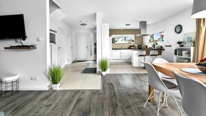 A modern kitchen with white cabinets, a dining area with a wooden table and chairs, and a TV mounted on the wall.