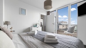 A modern bedroom with a large bed, a bedside table with a lamp, a small plant, and a view of the outside balcony.