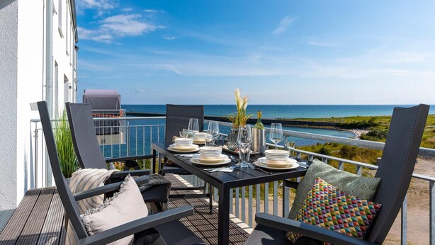 Une table à manger dressée pour six personnes, avec vue sur la mer, des sièges d’extérieur et une rambarde.