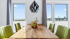 Un coin repas avec une table en bois, des chaises à l’assise rembourrée de tissu vert, un luminaire suspendu et une vue sur le paysage grâce à de grandes fenêtres.