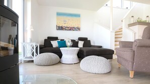 A modern living room with a black sofa, white pouf, and a glass coffee table. A painting of a beach scene hangs on the wall.