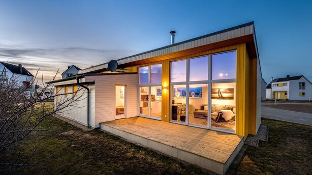 A modern house with large glass windows, a wooden deck, and a visible kitchen area.