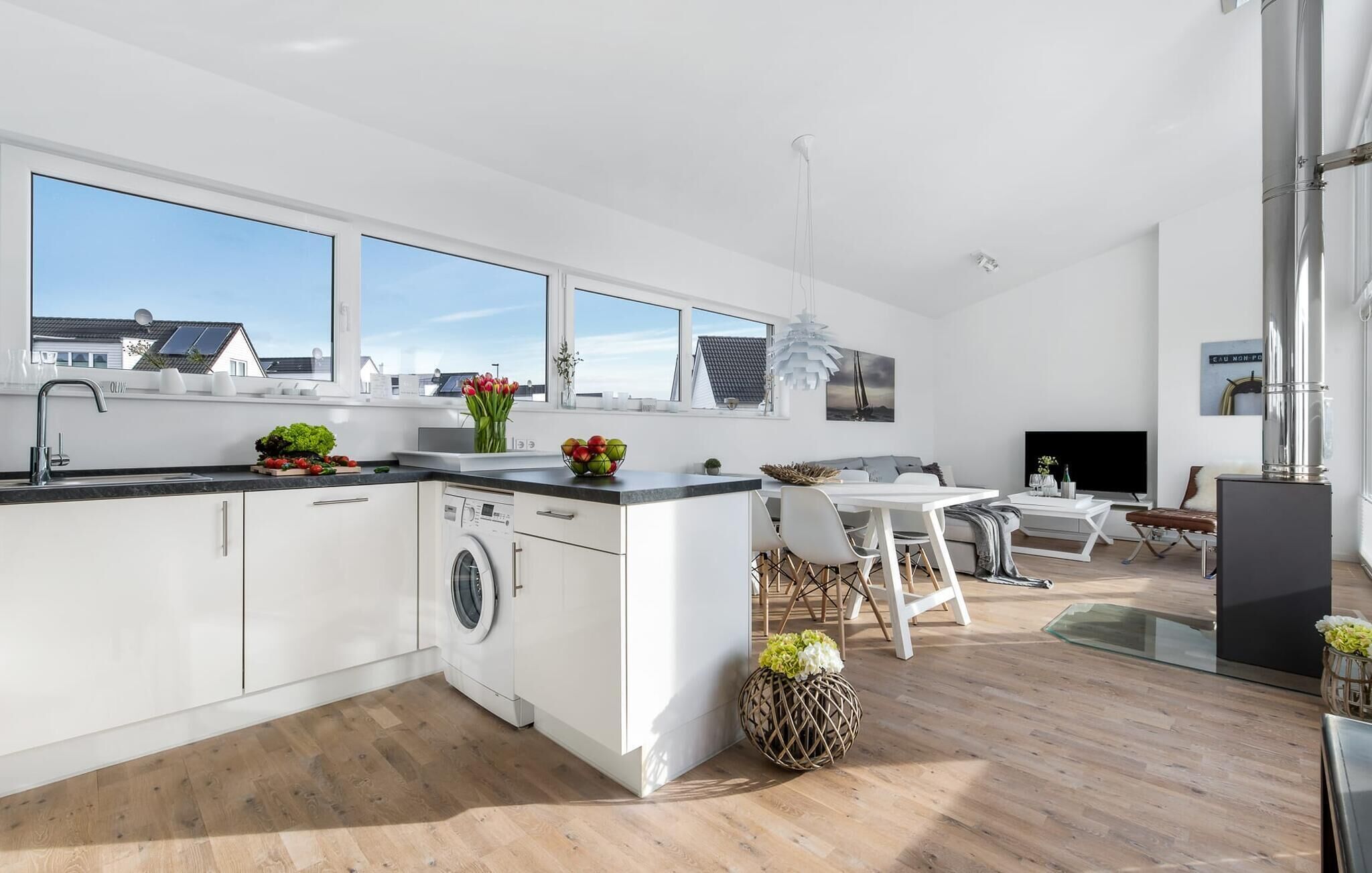 A modern kitchen with a built-in washing machine, a dining area with a white table and chairs, and a large window offering a view of the city.
