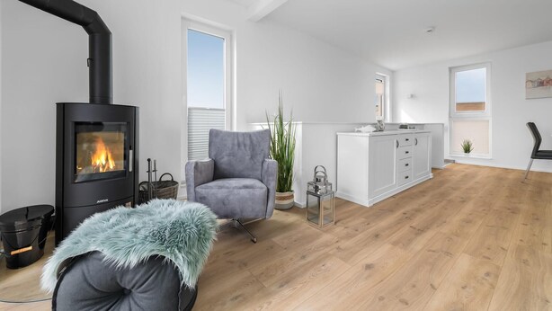 A modern living room with a wood-burning stove, a grey armchair, a small table with a plant, and a kitchen area with white cabinets. - Lovely apartment in Olpenitz with sauna (Olpenitz)