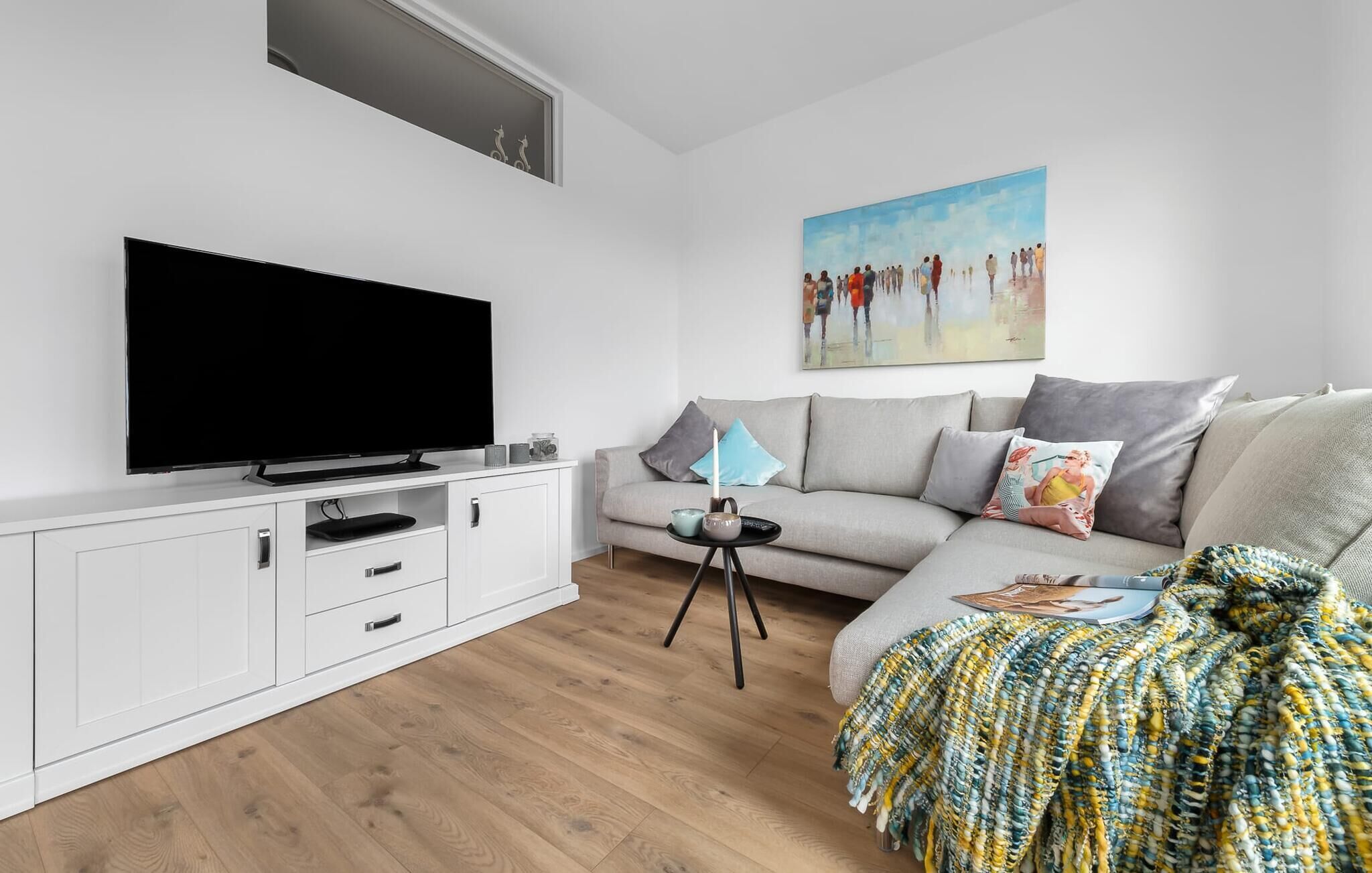 Ein modernes Wohnzimmer mit einem Flachbildfernseher, einem grauen Sofa, einer gemusterten Decke und einem Bild an der Wand.