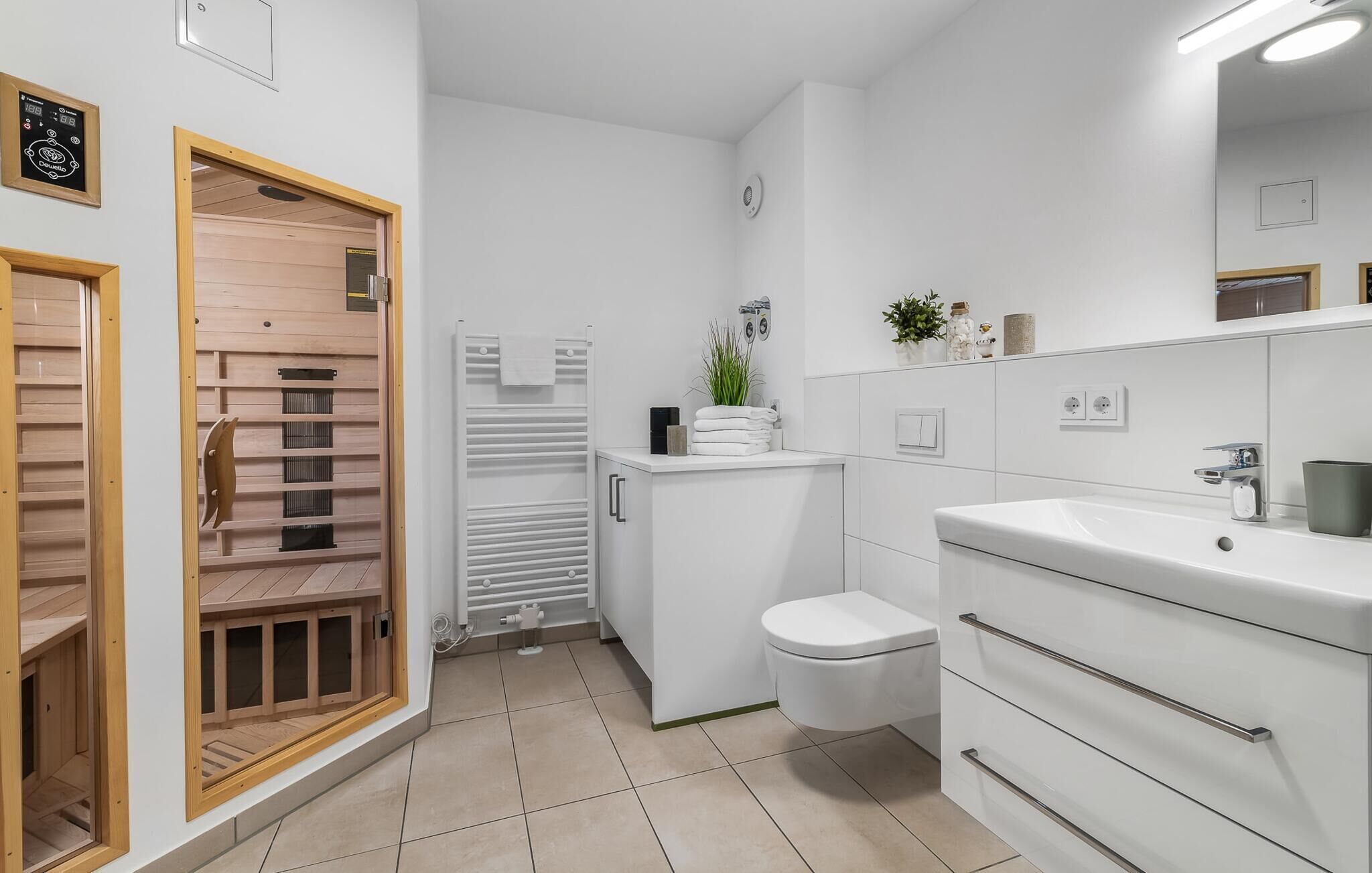 Un baño moderno con inodoro, lavabo y zona de ducha. Se aprecia una puerta de madera con ventana, un radiador blanco y una planta pequeña sobre el lavabo.