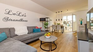 A modern living room with a grey sofa, a round coffee table with fruit, a dining area with chairs, and a kitchen area with a fridge.