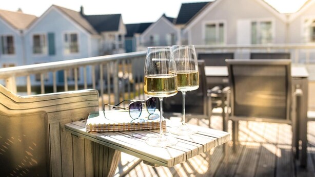 A wooden deck with two glasses of white wine, a notebook, and a pair of sunglasses.
