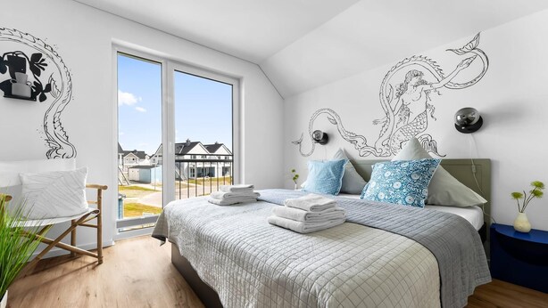 Une chambre moderne avec un grand lit, une vue sur les maisons à travers une porte coulissante et des œuvres d’art décoratives au mur.