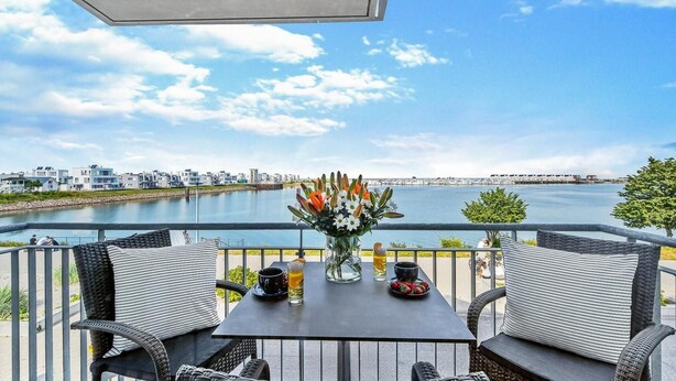 A balcony with wicker chairs, a table with drinks and a flower vase, overlooking a waterfront with buildings and a pier. - Pet friendly apartment in Olpenitz (Olpenitz)