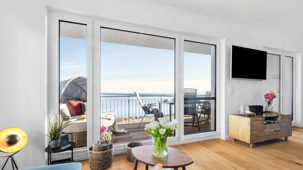 Un salon moderne doté d’une grande porte-fenêtre coulissante donnant sur un balcon avec vue sur la mer. On y trouve un téléviseur fixé au mur, un meuble en bois et une petite table avec des boissons.