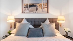 Une chambre avec un grand lit, deux lampes de chevet, un tableau encadré représentant une plage et un vase rempli de fleurs sur la table de chevet.