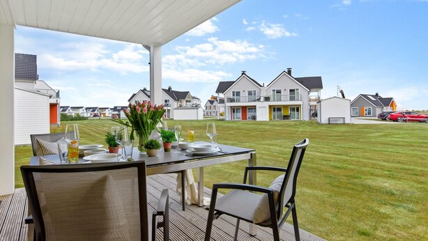 A covered outdoor dining area with a table set for four, surrounded by chairs, overlooking a grassy lawn and residential buildings.