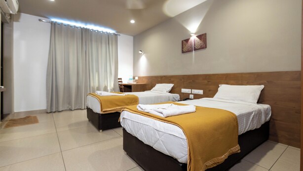 A hotel room with two beds, a wooden headboard, a small bedside table, and a curtain. - Aryaas Residency Nettoor (Kanayannur)
