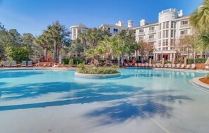 A large pool with a circular island, surrounded by palm trees and lounge chairs, with a grand building in the background. - Beautiful 1-bedroom condo with WiFi, at Sandestin Resort - Elation Building (Miramar Beach)