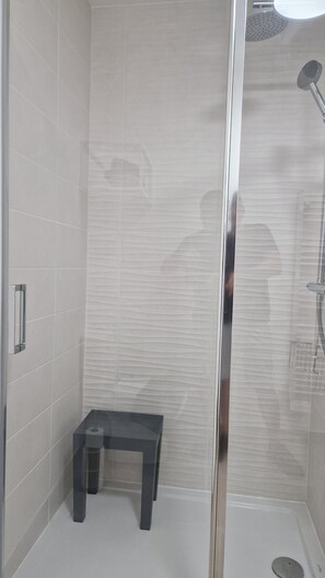 A modern shower area with a glass door, a small bench, and a showerhead. - Archipelago terrace side (Valras-Plage)