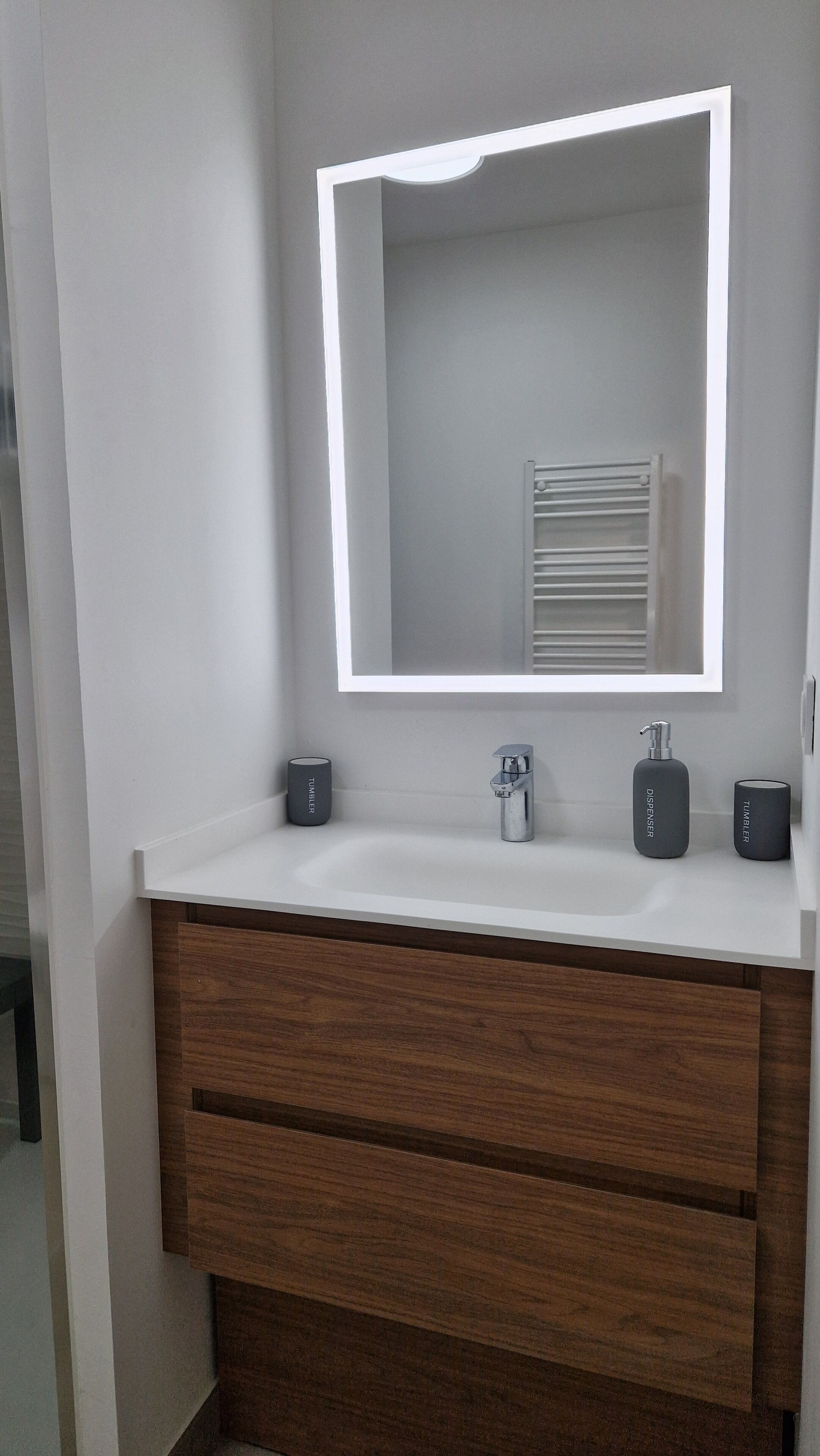 Un meuble-lavabo de salle de bain avec un plan de toilette blanc, un miroir doté d’un éclairage intégré, un robinet et deux distributeurs portant l’inscription « TUMBLER ».