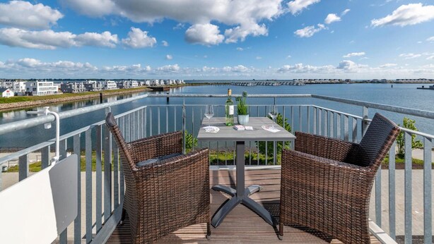 Un balcon avec des fauteuils en osier, une table et une bouteille de vin, donnant sur une étendue d’eau et un port de plaisance.