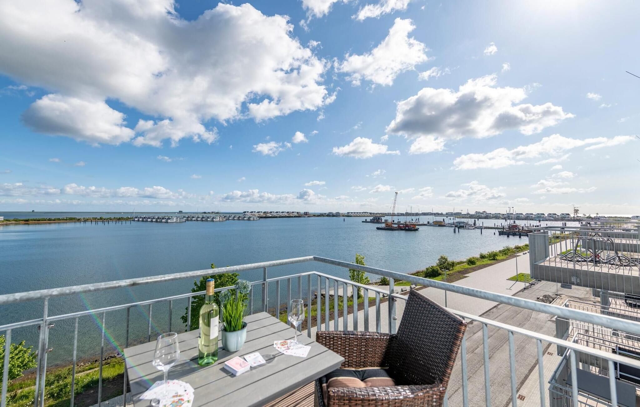 A balcony with a view of a marina, a dining table with a bottle and glass, and a wicker chair.