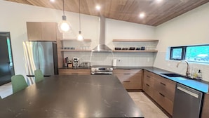 A modern kitchen with a large island, wooden ceiling, pendant lights, and a view through a window. - Spruce House: The Perfect Summer Retreat with Hiking, Swimming, and More (Copper Harbor)