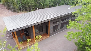 A modern two-story building with a metal roof, a covered porch with seating, and large windows. - Spruce House: The Perfect Summer Retreat with Hiking, Swimming, and More (Copper Harbor)